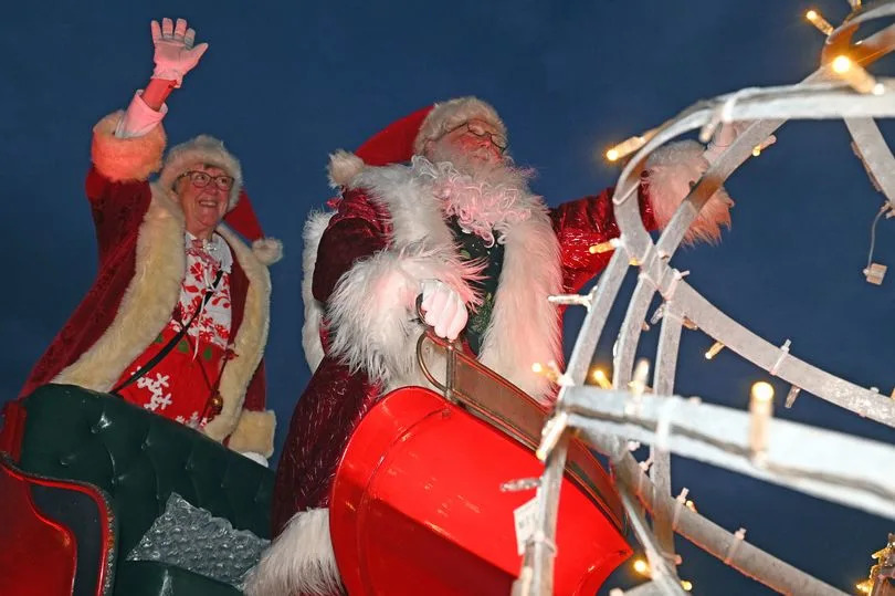 A person dressed as Santa Claus. He is dressed holding a metal rail, and woman in Christmas reds is standing behind him waving.