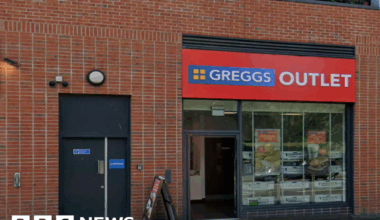 The outside of a Greggs outlet. It has the usual logo on the sign - four orange squares, in a square, on a blue background - but is surrounded by a red border with "outlet" written on it in white letters.