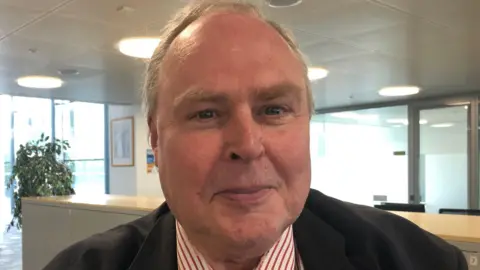 Vikki Irwin/BBC Richard Smith facing the camera and smiling. He is wearing a black suit with a red and white striped shirt underneath. Behind him is an office-style room. 