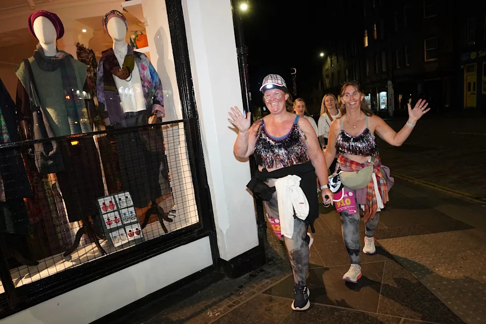 Women in sparkly bras wave at the camera as they walk past a clothes shop in Edinburgh during this year's MoonWalk event