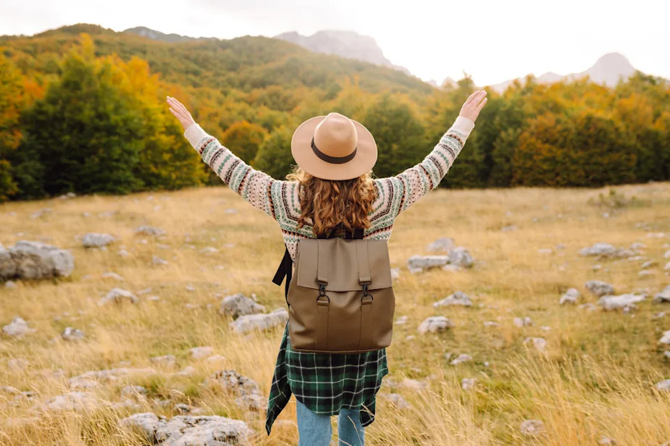 Person with hat and backpack stands in a grassy field with arms raised, surrounded by trees and mountains in the distance