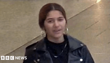 Pantica Sali wearing a black leather jacket over a black top with a chain around her neck. She has hooped earrings and black hair tied back.