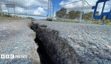 A large crack is running through the middle of a Tarmac road. There barriers across the road to close it off to traffic.