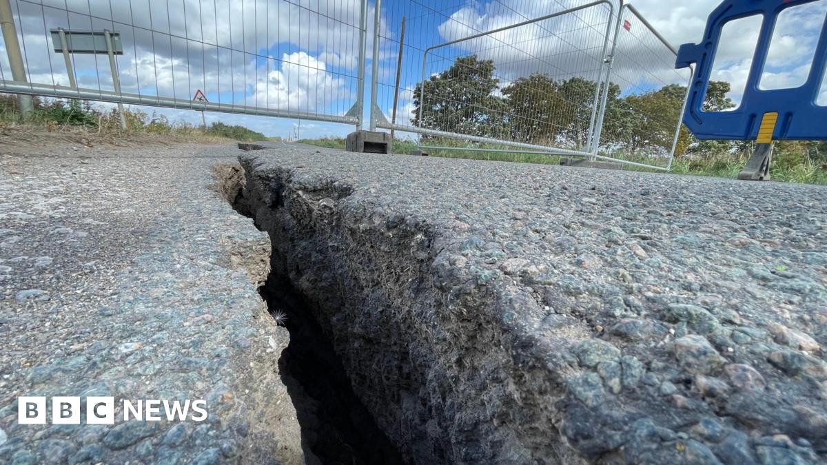 A large crack is running through the middle of a Tarmac road. There barriers across the road to close it off to traffic.