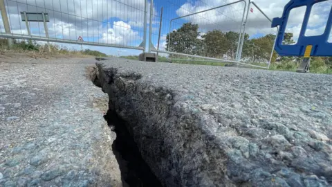 BBC A large crack is running through the middle of a Tarmac road. There barriers across the road to close it off to traffic. 