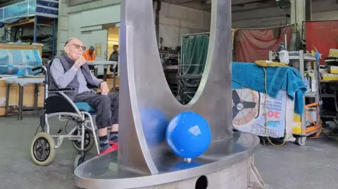 Benson-Sedgwick Engineering Ltd An elderly man sits in a wheelchair next to the large steel structure in a busy warehouse.