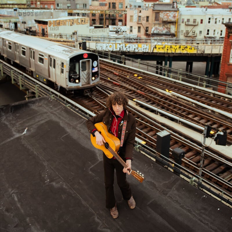A man with curly hair plays an acoustic guitar on a rooftop overlooking subway tracks, with a silver train passing by and graffiti-covered buildings in the background.
