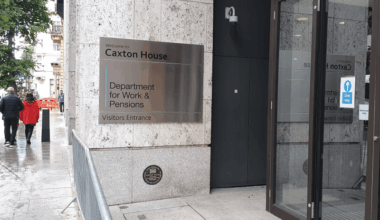 A silver sign on a wall says Welcome to Caxton House, Department for Work and Pensions, Visitors Entrance, with people walking away from the camera on the pavement to the left
