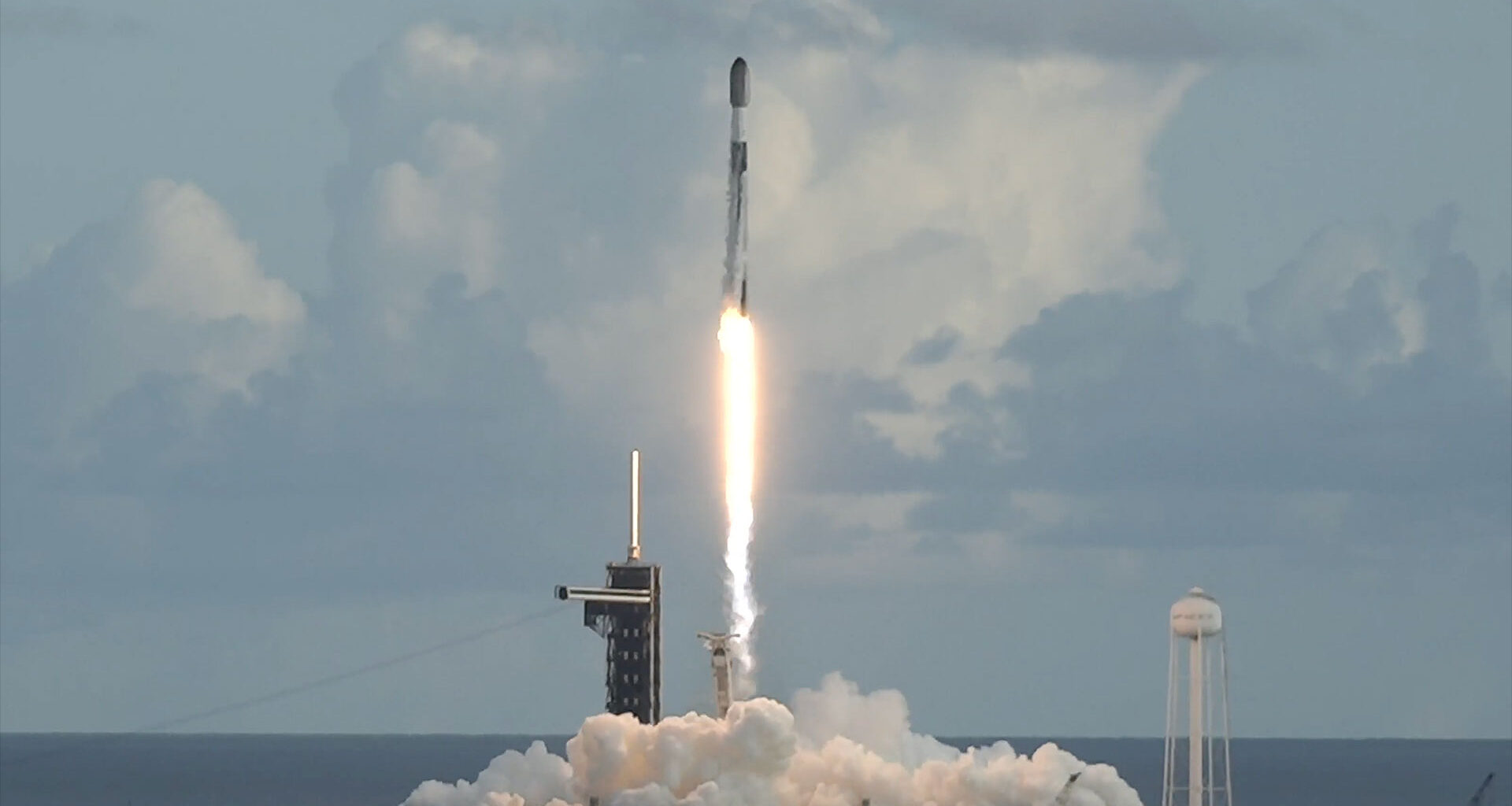A white and black rocket lifts off into a partially cloudy sky above the East Coast of the United States, riding atop the bright white exhaust of its nine engines.