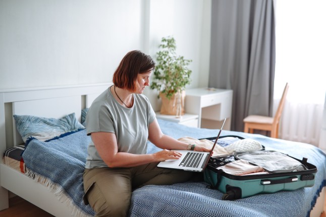 A woman sitting on a bed using a laptop, next to her open packed suitcase.