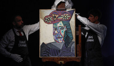 two men in aprons, white dress shirts, and white gloves presenting a colorful painting of a woman from the chest up