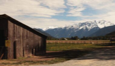 A rustic wooden barn stands on a grassy field with a backdrop of snow-capped mountains under a partly cloudy sky. A dirt path runs alongside the barn.