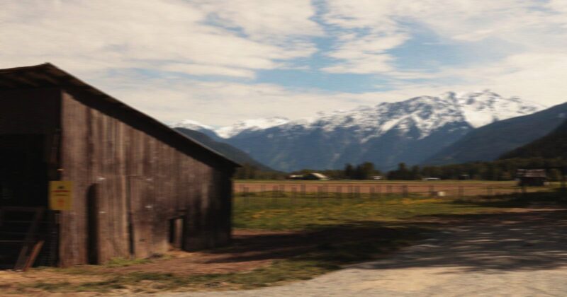 A rustic wooden barn stands on a grassy field with a backdrop of snow-capped mountains under a partly cloudy sky. A dirt path runs alongside the barn.