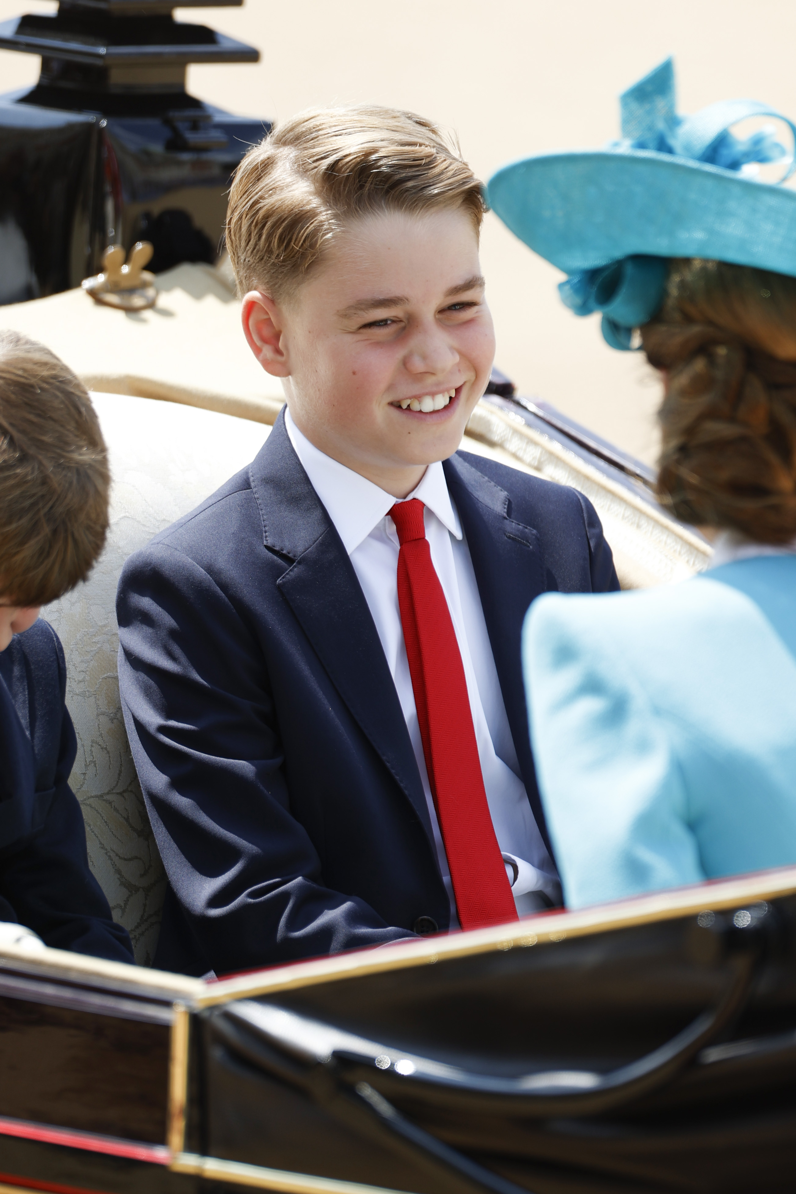 Prince George riding in a carriage in a blue suit and red tie