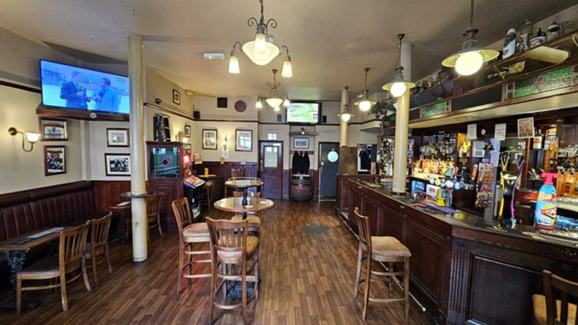 Interior of a pub with a long wooden bar, several tables and chairs, and a television playing sports.