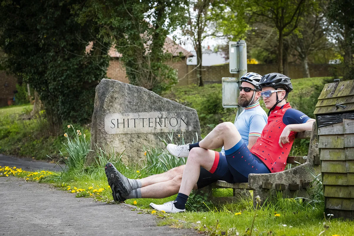 Two cyclists sitting next to stone sign for the hamlet of 'Shitterton'