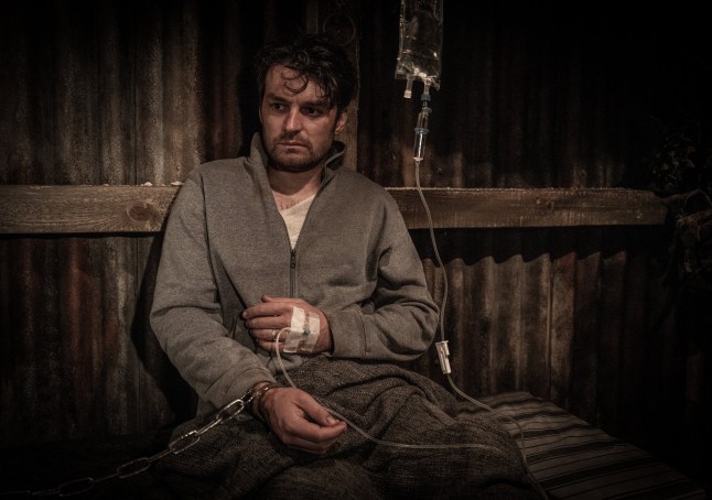 Emmerdale's Mackenzie Boyd being held captive in an underground bunker, with a chain on his wrist and a cannula in his hand. His clothes are grubby and he is under a blanket