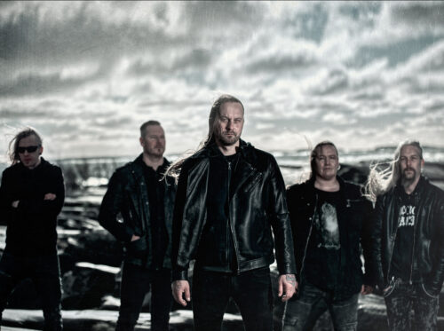 Band photo of Mors Principium Est (2025), five members in black leather standing on a rocky shore under a cloudy sky.