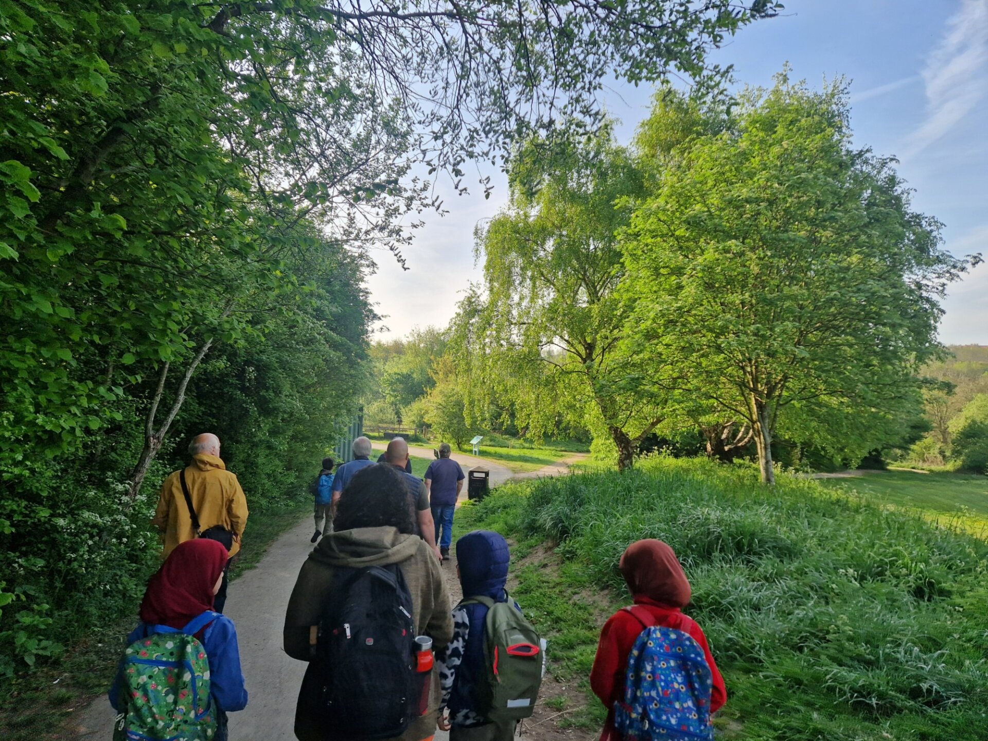 A group of adults and children are walking away from the camera along a footpath. There are big green trees, grass and hedges all around them. It's a sunny day.