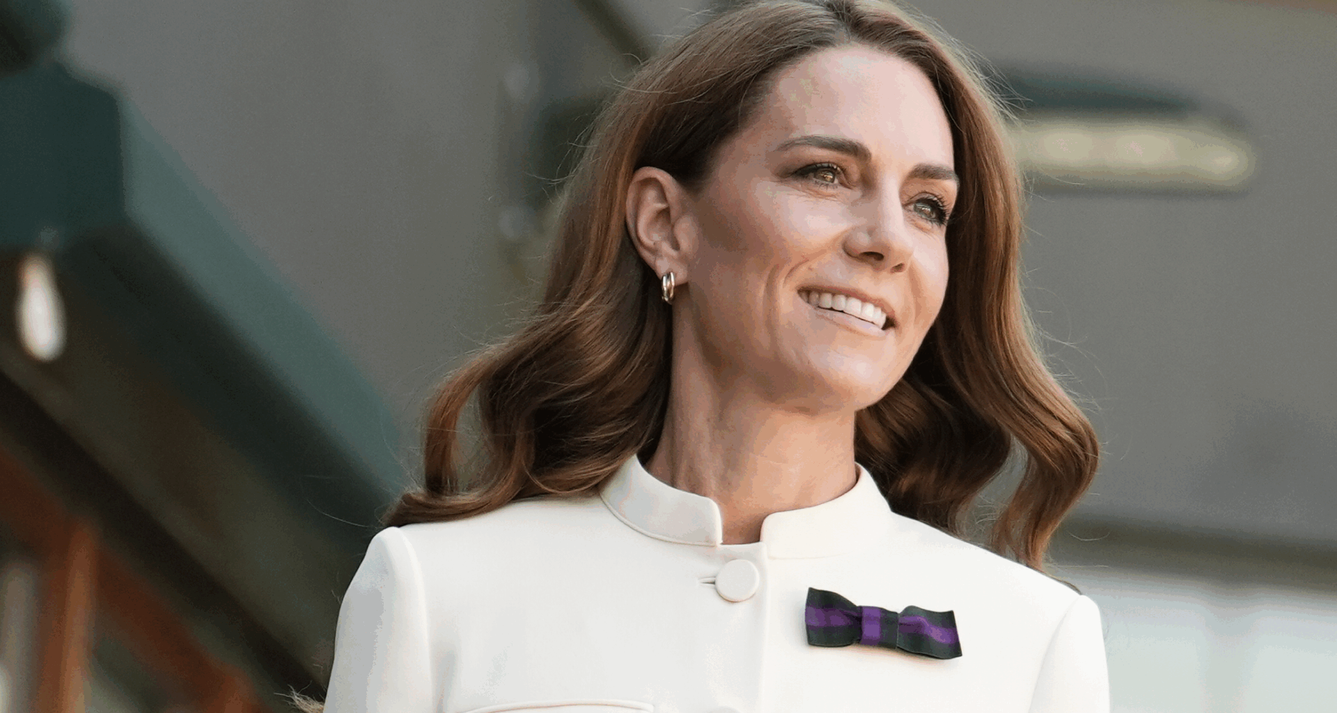 Kate Middleton wearing a white blazer with a purple bow pin and smiling