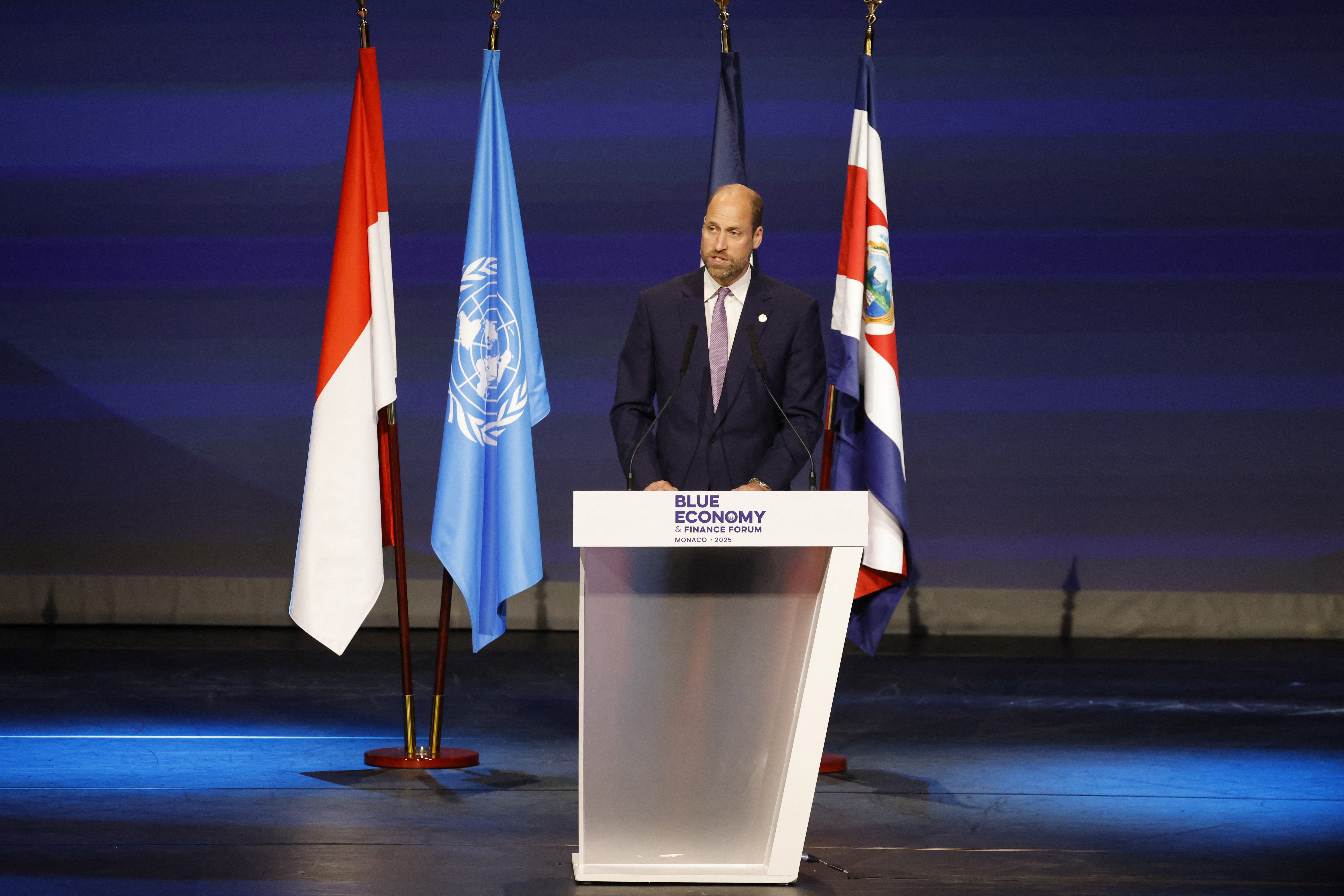 Prince William giving a speech in front of four flags on a stage