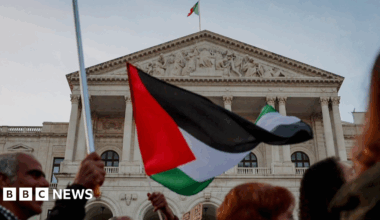 Blurred heads of people walking across the front/bottom of the screen with one of them holding a Palestinian flag which is centre and focus of the image in front of the Portuguese parliament in Lisbon which is a six columned grand neoclassical building with a white marble facade and a Portuguese flag on top