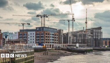 Several cranes stand over riverside construction sites, where new apartment blocks and modern buildings are being built. In the foreground, a moored barge rests on the riverbank beside the water, while scaffolding and partially completed structures dominate the skyline under a cloudy sky.