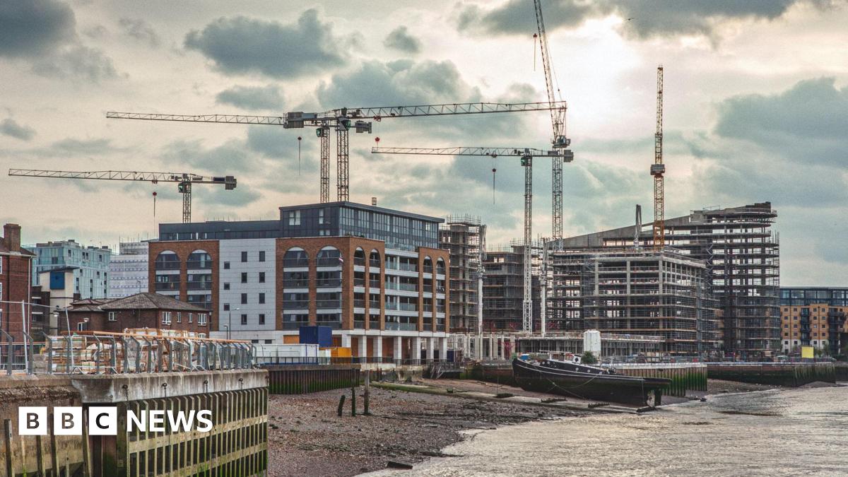 Several cranes stand over riverside construction sites, where new apartment blocks and modern buildings are being built. In the foreground, a moored barge rests on the riverbank beside the water, while scaffolding and partially completed structures dominate the skyline under a cloudy sky.