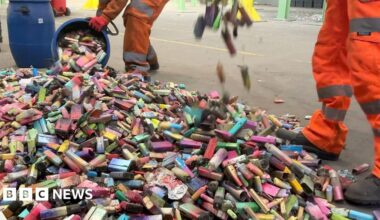 A large pile of dirty used vapes on the concrete floor of a recycling facility. The vapes are in different shapes, sizes and colours. Two workers in orange overalls are adding to the pile from blue bins.