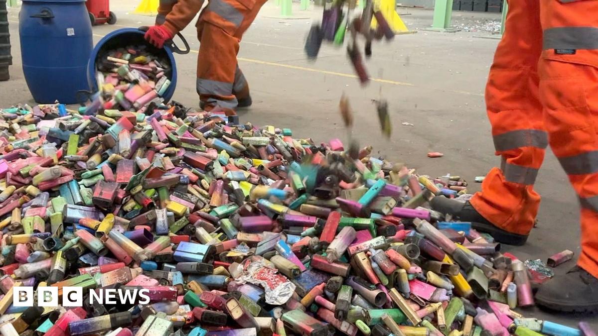 A large pile of dirty used vapes on the concrete floor of a recycling facility. The vapes are in different shapes, sizes and colours. Two workers in orange overalls are adding to the pile from blue bins.