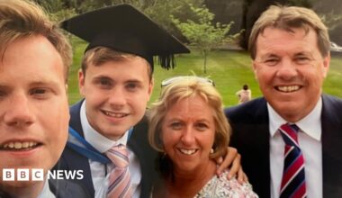 Jack O'Sullivan, pictured on his graduation day with his family.  His brother is on the left of the frame. He has fair short hair and is smiling. Jack is next to him. He also has short fair hair and is wearing a graduation cap and gown over a white shirt and pink and blue striped tie. Catherine is next to him. She has short blonde hair and has glasses on her head. She is looking at the camera and smiling. Alan O'Sullivan is on the right and is wearing a suit.