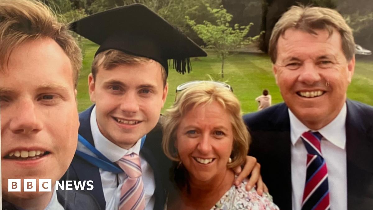 Jack O'Sullivan, pictured on his graduation day with his family.  His brother is on the left of the frame. He has fair short hair and is smiling. Jack is next to him. He also has short fair hair and is wearing a graduation cap and gown over a white shirt and pink and blue striped tie. Catherine is next to him. She has short blonde hair and has glasses on her head. She is looking at the camera and smiling. Alan O'Sullivan is on the right and is wearing a suit.