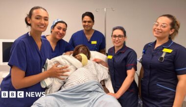 Staff using the simulation lab are standing around Linda the mannequin who has just given birth. The maternity staff are wearing blue uniforms with yellow name badges and holding a doll baby wrapped in a blanket and with a knitted yellow hat on. There is equipment around them, including a screen.