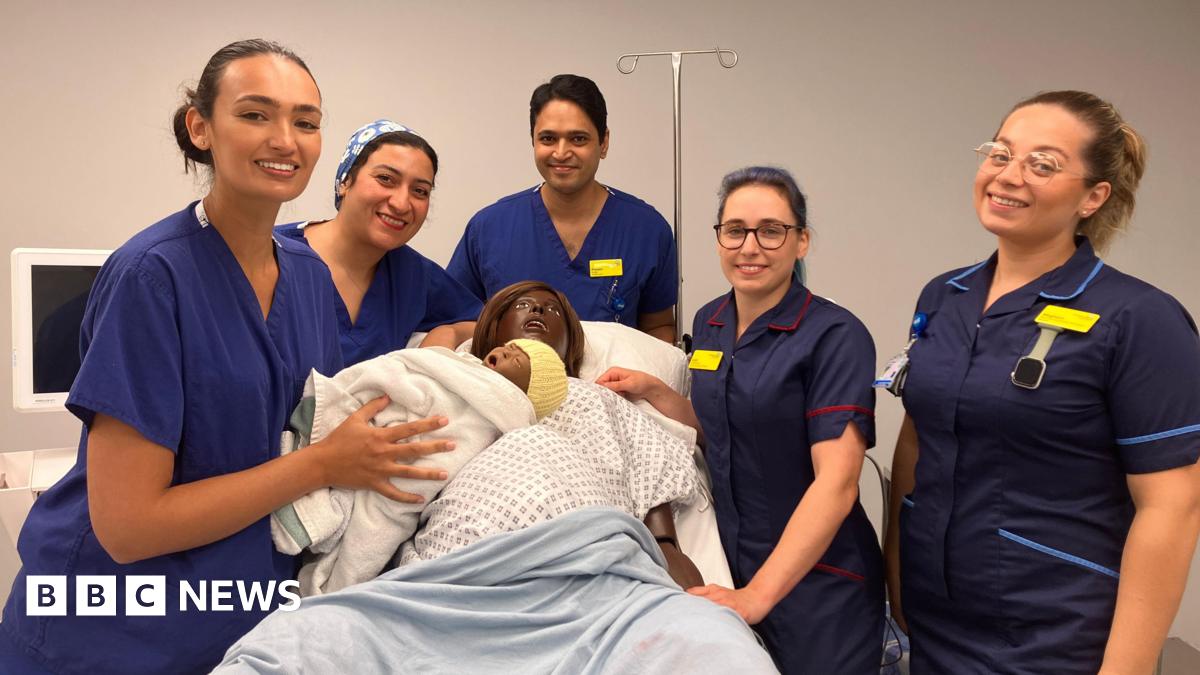 Staff using the simulation lab are standing around Linda the mannequin who has just given birth. The maternity staff are wearing blue uniforms with yellow name badges and holding a doll baby wrapped in a blanket and with a knitted yellow hat on. There is equipment around them, including a screen.
