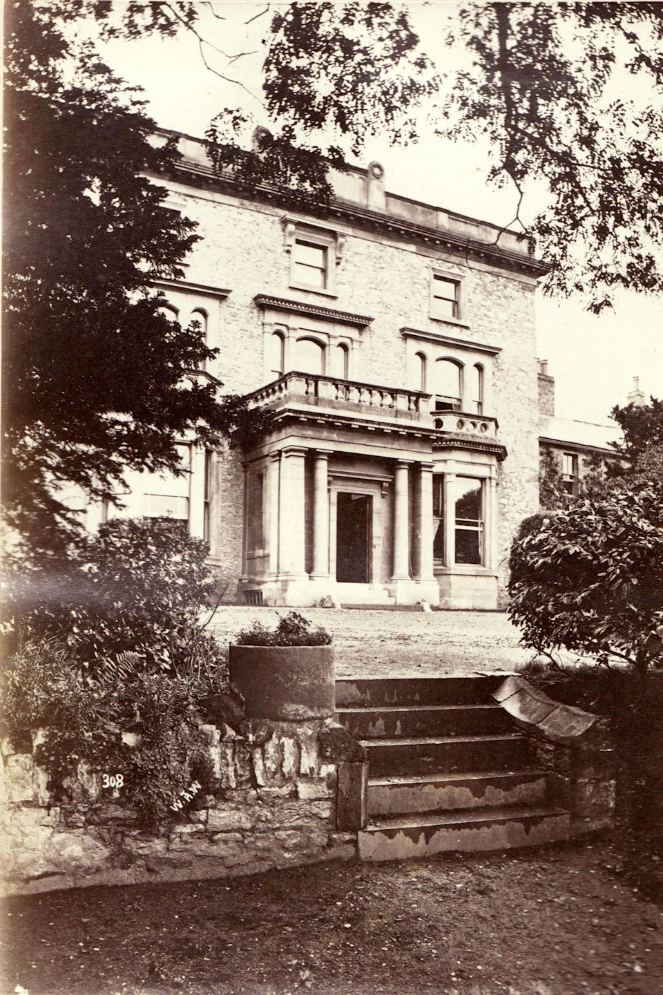 The driveway leads to a porticoed entrance before a Georgian-style manor house.  Steps lead down to a garden area.