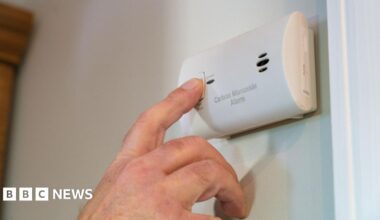 A close-up of a person pressing the test button on a carbon monoxide alarm mounted on a wall.