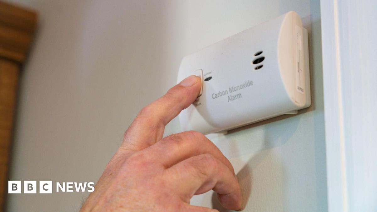 A close-up of a person pressing the test button on a carbon monoxide alarm mounted on a wall.