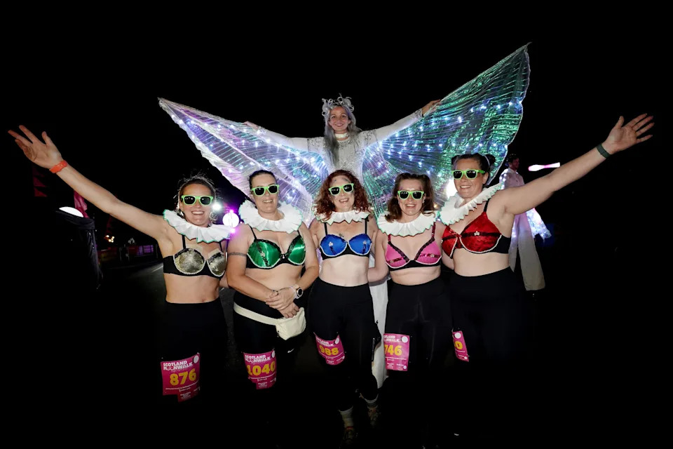A group of women in multi-coloured bras with another woman behind them dressed like an angel or butterfly with illuminated wings