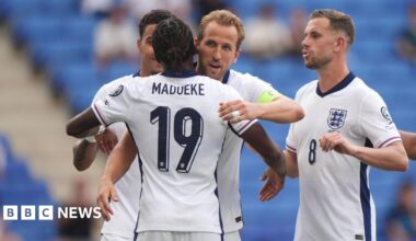 Harry Kane is about to hug his teammate, Noni Madueke. They are wearing white England football shirts with navy blue accents on them. Harry Kane is wearing a bright green captain's armband. Next to Harry is Jordan Henderson, moving in for a hug.