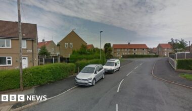 An asphalt road with white line markings leads to a series of council house. Two cars, one white, the other silver are parked straddling the road and pavement.