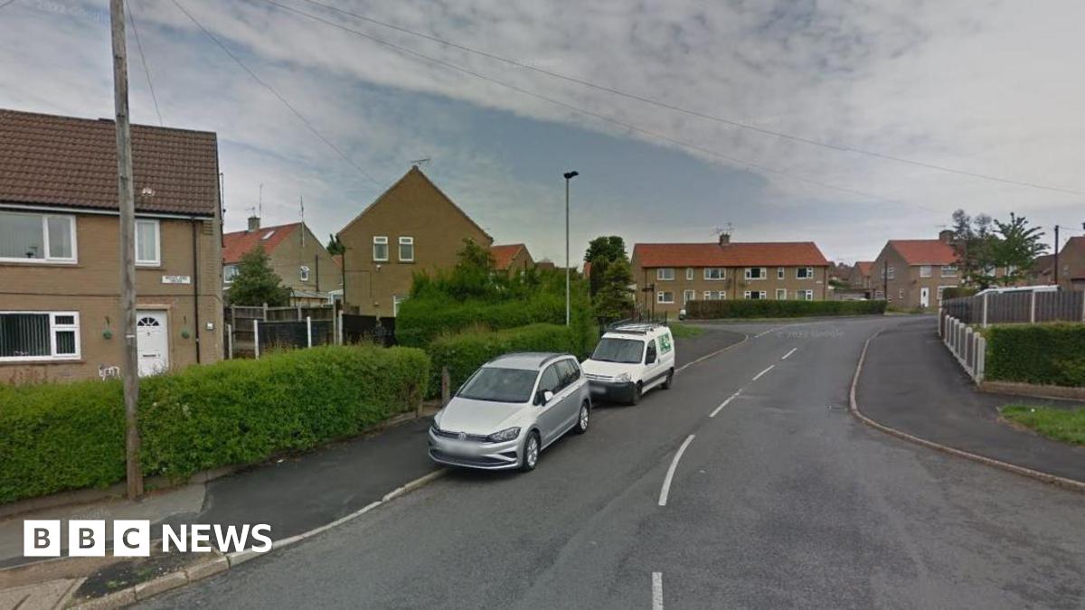 An asphalt road with white line markings leads to a series of council house. Two cars, one white, the other silver are parked straddling the road and pavement.