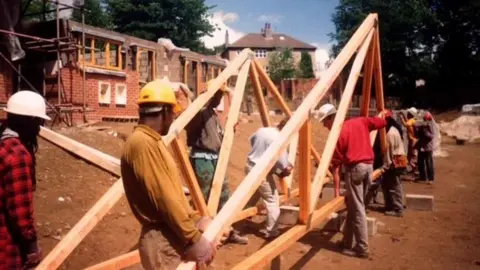 Frontline Self-Build An old image of people carrying a section of roofing in Leeds. In the background is a housing construction site. The people are wearing hard hats.