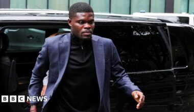 Thomas Partey wears a navy blue blazer and black shirt. He walks from a black car towards the camera.