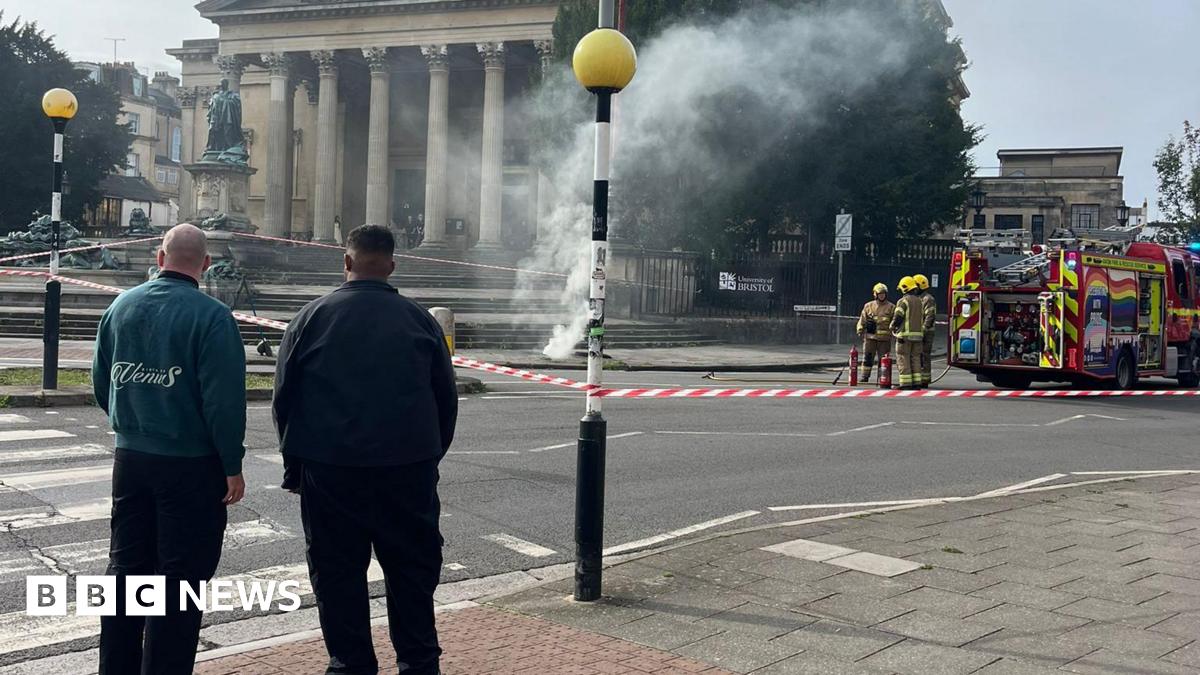 Two men stand and look across a road, in front of a zebra crossing. On the right is a fire engine. In the centre smoke pours from the pavement.