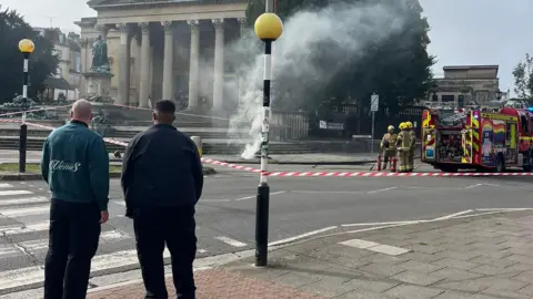 BBC Two men stand and look across a road, in front of a zebra crossing. On the right is a fire engine. In the centre smoke pours from the pavement.