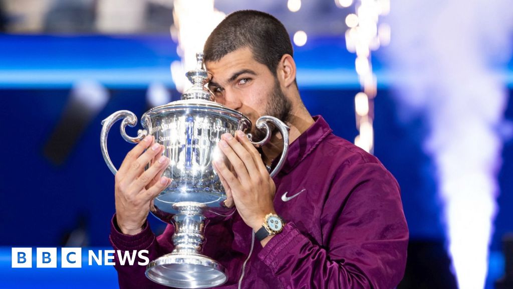 Carlos Alcaraz, wearing a purple Nike jacket, kissing the US Open trophy.