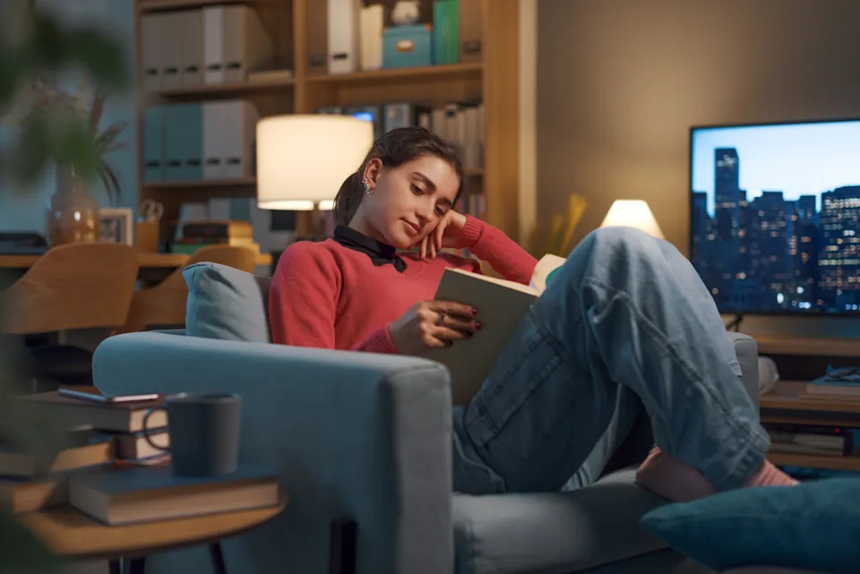 Person relaxing on a couch reading a book in a cozy room with a cityscape visible on a TV screen in the background