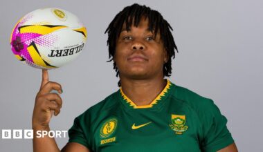 Babalwa Latsha, wearing a green South Africa rugby shirt in front of a grey background, looks directly at the camera as she balances a rugby ball on her left index finger