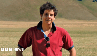 A boy with dark curly hair in a red polo shit stands, smiling at the camera, with his hands on his hips, in front of a field and hills
