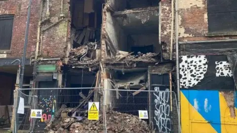 Cara Thorpe/BBC Metal fences sit at the feet of a collapsed building. Fallen brickwork and debris can be seen behind.